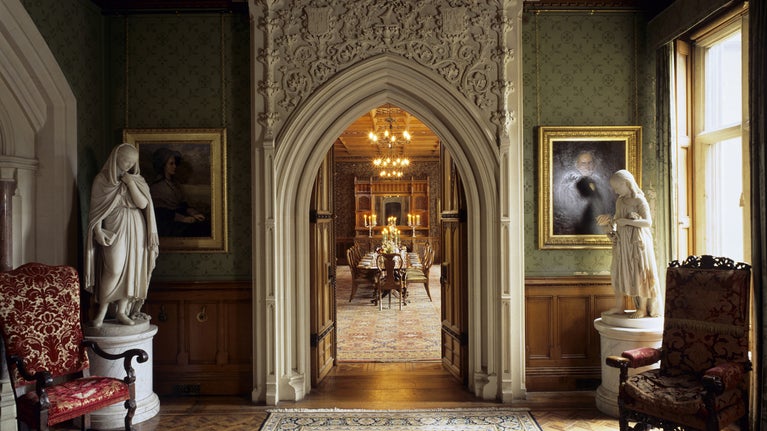 View of Dining Room from the Oak Lobby at Tyntesfield with table laid, candles & chandelier lighted and statue of Albinia Gibbs (1863 Rome) one side of stone doorway.